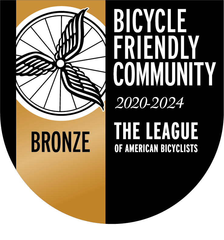 A seal in the shape of a shield reads “BICYCLE FRIENDLY COMMUNITY 2020-2024 THE LEAGUE OF AMERICAN BICYCLISTS” on a black background, with the League’s seal placed over a bronze vertical stripe, with the word “BRONZE” below the seal.