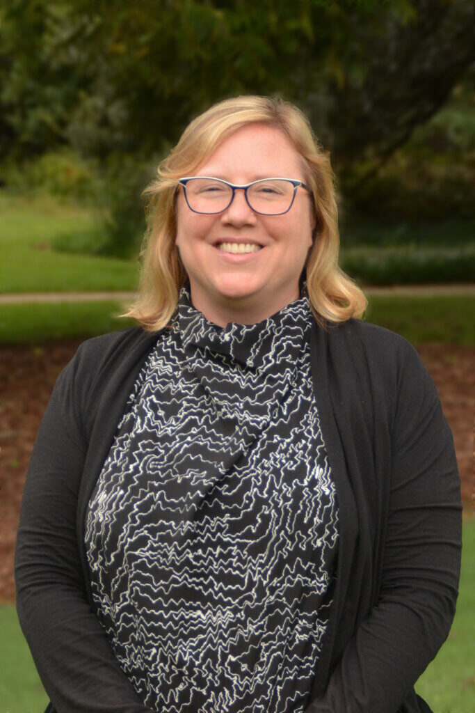 Photo of a blond woman wearing a black and white shirt and a black sweater. The woman is smiling and has glasses, and is standing in front of a green space.