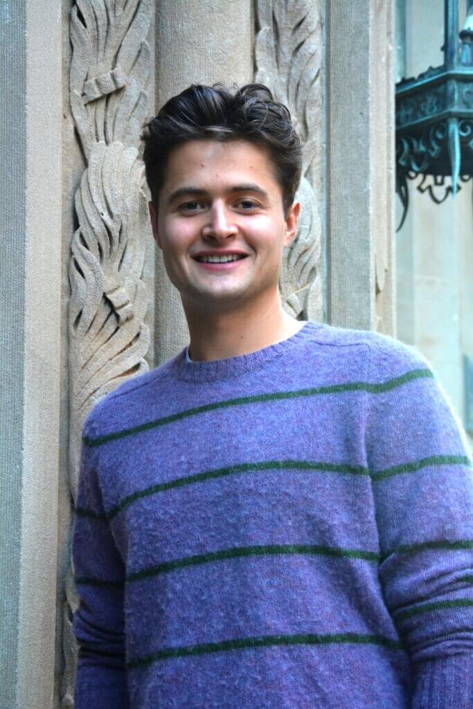 Photograph of Tristan Kercher, in a purple sweater with grey stripes, standing against a historic Main Street building.