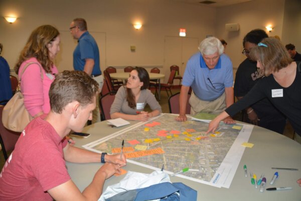 A photo of individuals gathered around a table with maps at the design workshop, some are writing on sticky notes and some are pointing at the map.