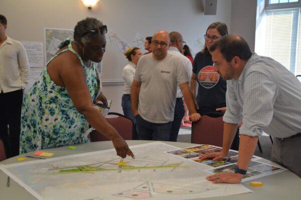 A photo of individuals gathered around multiple tables with different conceptual designs at the second day of the design workshop.