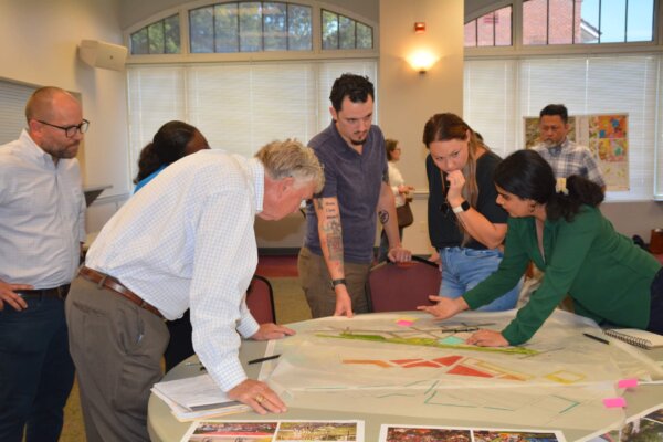 A photo of individuals gathered around multiple tables with different conceptual designs at the second day of the design workshop.
