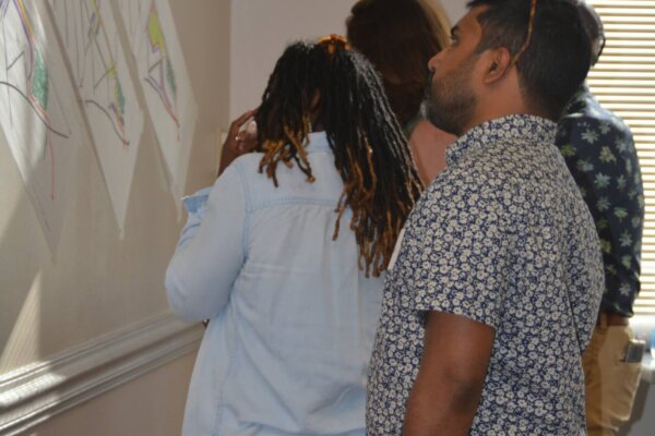Individuals stand and review the early concepts explored in response to day 1 of the design workshop, which are taped to the wall