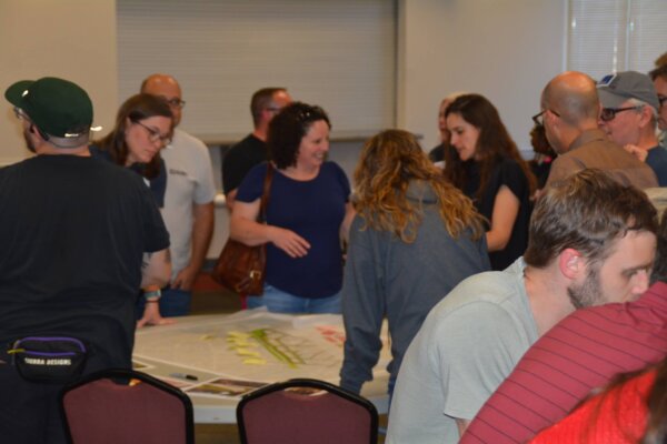 A photo of individuals gathered around multiple tables with different conceptual designs at the second day of the design workshop.
