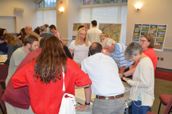 A photo of individuals gathered around multiple tables with different conceptual designs at the second day of the design workshop.