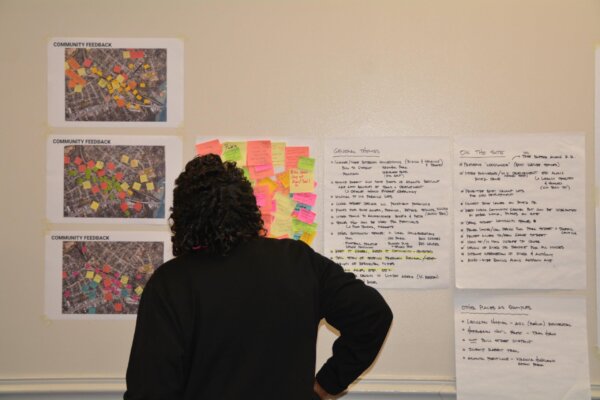 A photo of a woman standing and reviewing the feedback received and general themes that emerged as presented on the wall via printouts, sticky notes, and an outline