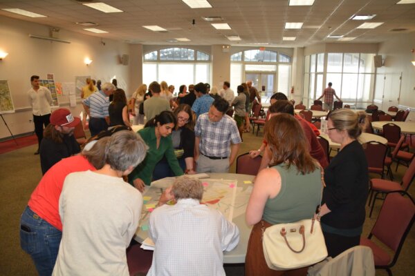 A photo of individuals gathered around multiple tables with different conceptual designs at the second day of the design workshop.