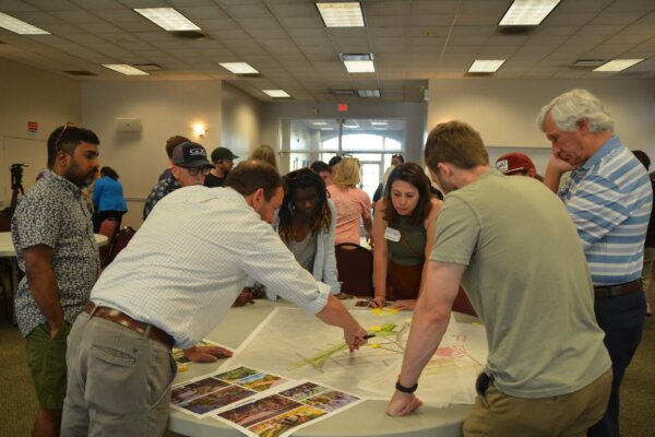 A photo of individuals gathered around multiple tables with different conceptual designs at the second day of the design workshop.