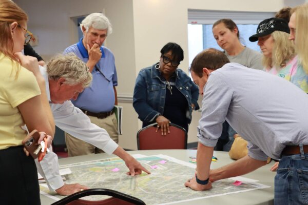 A photo of individuals gathered around a table with maps at the design workshop; some are pointing at the map on the table.