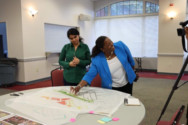 A photo of individuals gathered around multiple tables with different conceptual designs at the second day of the design workshop.