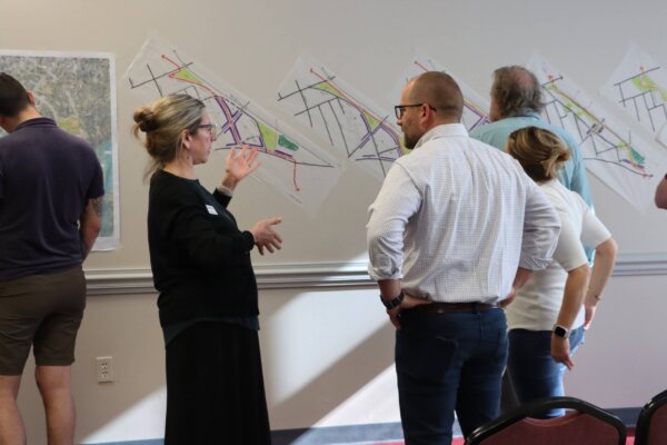 Individuals stand and review the early concepts explored in response to day 1 of the design workshop, which are taped to the wall