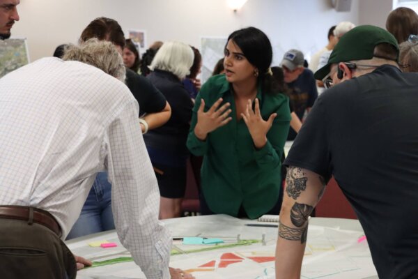 A photo of individuals gathered around multiple tables with different conceptual designs at the second day of the design workshop.