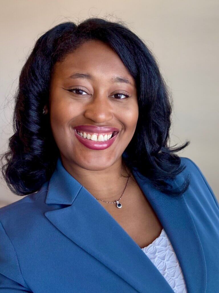 Head shot of Nicole Broner, who is wearing a blue suit and standing against a beige background